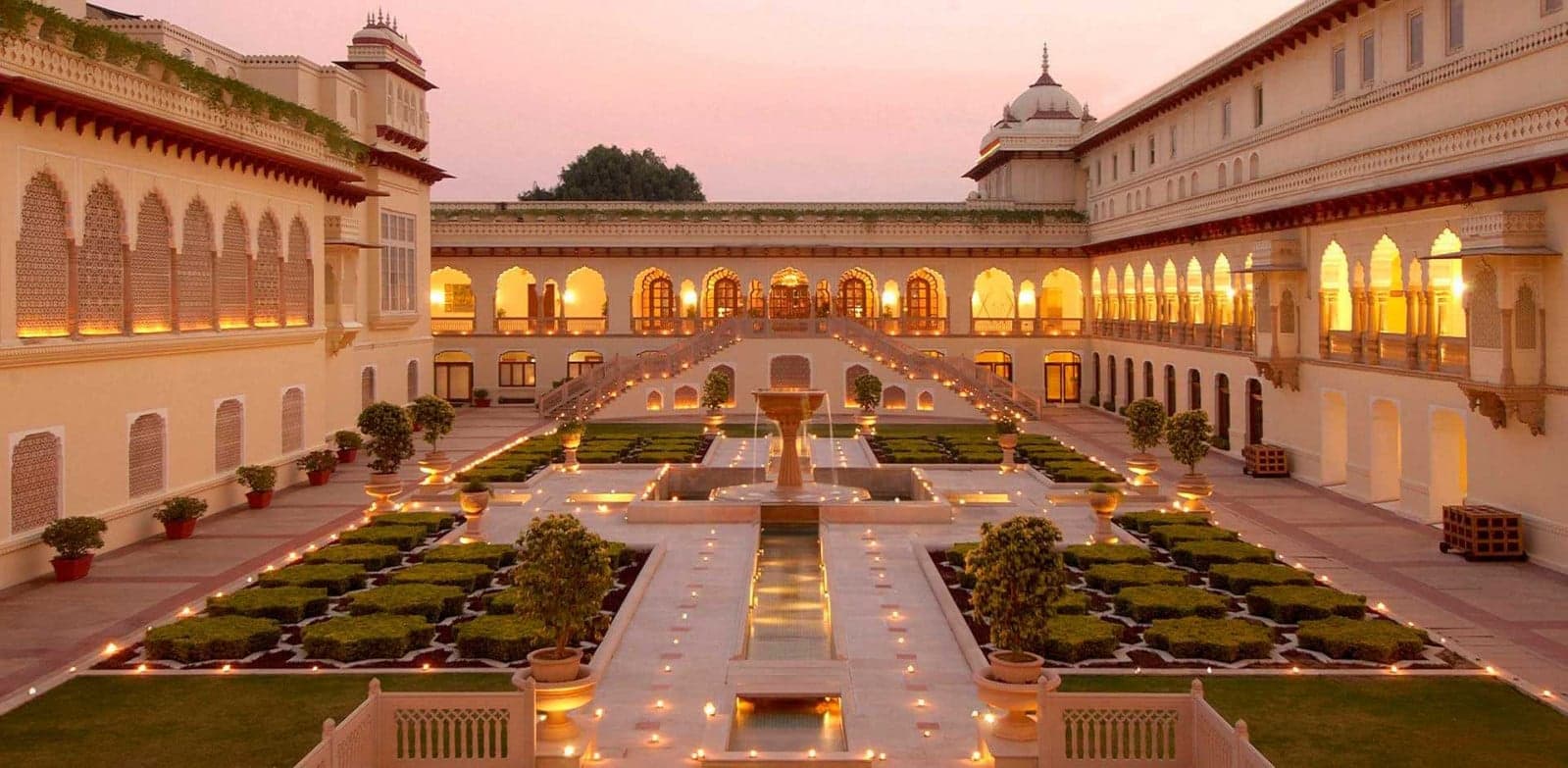 An illuminated palace garden courtyard with fountains, hedges and arched colonnades at dusk