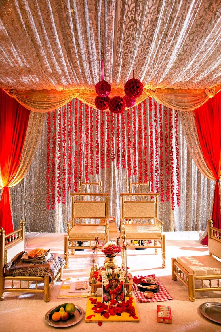 An ornate wedding mandap draped in red and gold fabrics with hanging floral garlands